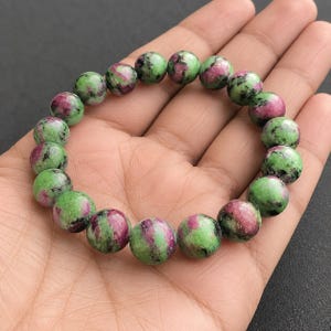 Könnte beinhalten: Ein Perlenarmband aus runden, polierten Steinen mit einem gesprenkelten Muster in leuchtendem Grün und tiefem Magentarosa. Die Perlen sind in einer kreisförmigen Formation aufgereiht und zeigen natürliche Farbvariationen und Texturen.
