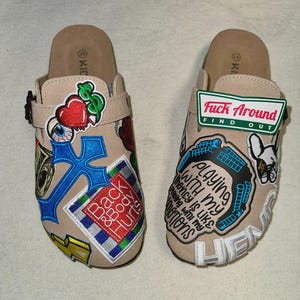 May include: A pair of beige clogs with various embroidered patches. One clog features a heart, eye, and cross design, while the other displays the text "Fuck Around Find Out" and other graphics. The clogs have a buckle strap.