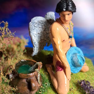 May include: A handcrafted figurine of a kneeling angel with black hair, wearing a chain necklace and holding a blue ball of yarn. The angel has white wings and is kneeling on a grassy surface with a brown pot.