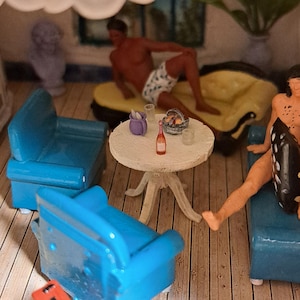 May include: Miniature scene of a leisure setting with a round table, blue armchairs, and a yellow sofa. A small bottle of wine, glasses, and a basket are on the table. Two figures are lounging, one on the sofa and the other in an armchair.