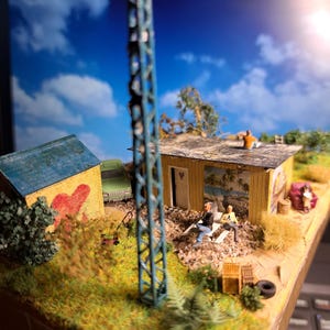 May include: Miniature diorama of a rural scene with a small house, a shed with a heart painted on it, and a tall blue tower. Two miniature figures are seated on lawn chairs. A green truck is parked nearby.