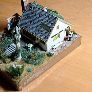 May include: Miniature model of a house with a gray tiled roof, surrounded by miniature landscaping including trees, bushes, and a small statue. The house has a white exterior with the words "Carpe diem" written on the side. A small figure stands near the door.
