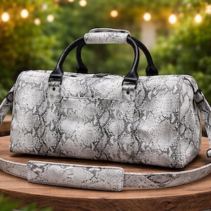 Puede incluir: Una bolsa de viaje gris y blanca con estampado de piel de serpiente, asas negras y una correa para el hombro. La bolsa está sobre una mesa de madera, con un fondo borroso de vegetación y luces. La bolsa tiene el texto "Streetrend Apparel".