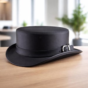 May include: A black leather top hat with a wide brim and a strap with a silver buckle. The hat is sitting on a wooden surface. The hat is a classic style.