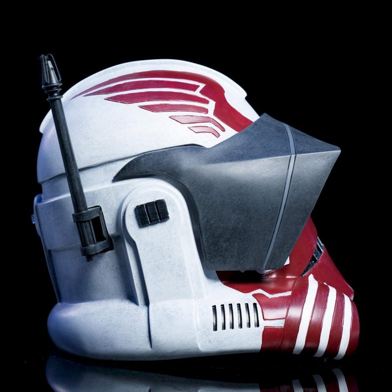 May include: A white and red helmet with a black visor and antenna. The helmet features red wing-like designs and vertical stripes. The design includes a series of vents and a rectangular button. The helmet is set against a black background.