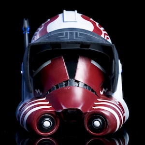 May include: A detailed, close-up view of a red, white, and black helmet. The helmet features a visor, two eye openings, and intricate white and red markings. The design is reminiscent of a science fiction or fantasy theme.
