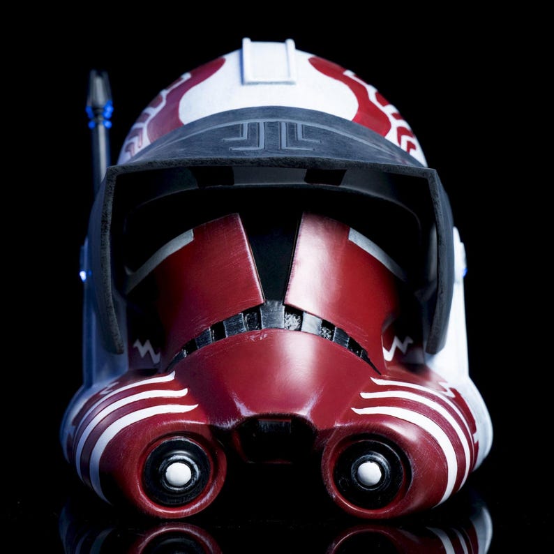 May include: A detailed, close-up view of a red, white, and black helmet. The helmet features a visor, two eye openings, and intricate white and red markings. The design is reminiscent of a science fiction or fantasy theme.