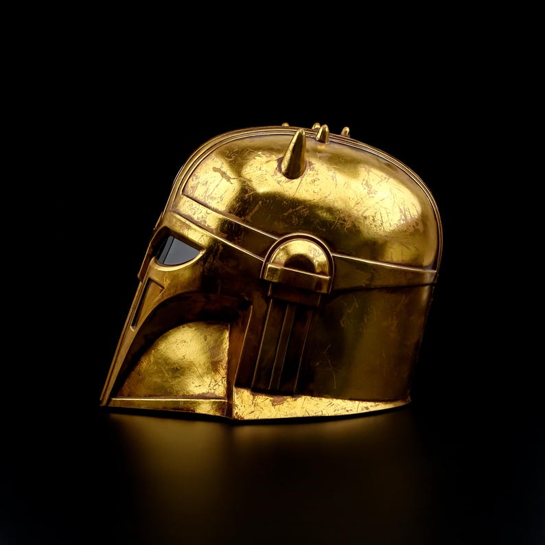 May include: A gold-colored helmet with a weathered finish, featuring a visor and distinctive angular design. The helmet has a series of small, pointed protrusions on top and is set against a black background. This item is likely a collectible or costume piece.