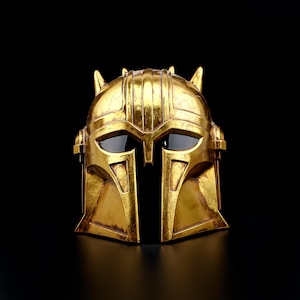 May include: A gold-colored helmet with a distinctive design, featuring a central vertical opening and dark eye coverings. The helmet has a textured surface and is set against a black background, highlighting its intricate details and metallic finish.