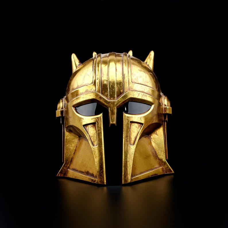 May include: A gold-colored helmet with a distinctive design, featuring a central vertical opening and dark eye coverings. The helmet has a textured surface and is set against a black background, highlighting its intricate details and metallic finish.