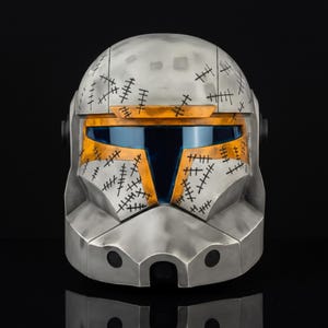 May include: A silver and orange helmet with a blue visor. The helmet has black stitch-like markings across the surface. The visor is framed with orange accents. The helmet is set against a black background.