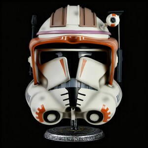 Puede incluir: Casco réplica detallado, principalmente blanquecino con detalles en naranja y marrón. El casco presenta una visera, dispositivos de comunicación y detalles intrincados, montado sobre un soporte decorativo. El diseño recuerda a un tema de ciencia ficción o fantasía.