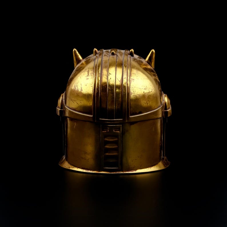 May include: A gold-colored helmet with a rounded top and vertical ridges. The helmet has two small, pointed protrusions on the top and a rectangular design on the front. The background is black.