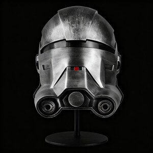 May include: A silver-coloured helmet with a distressed effect, featuring a small red square. The helmet is displayed on a black stand against a black backdrop. The design includes circular and angular details.