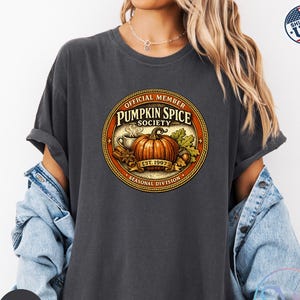 May include: A charcoal gray t-shirt featuring a circular graphic with the text Official Member Pumpkin Spice Society Est. 1997 Seasonal Division. The design includes a central orange pumpkin, a steaming mug, cinnamon sticks, and autumn leaves.