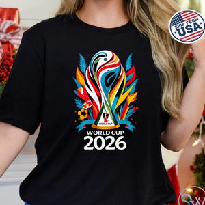 May include: A black short-sleeve t-shirt featuring a colorful graphic of the World Cup 2026 trophy. The design includes abstract shapes in red, blue, yellow, and green, with the text "WORLD CUP 2026" printed in white below the trophy illustration.