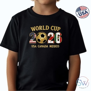 May include: Black t-shirt with a gold design featuring the text "WORLD CUP 2026" and a soccer ball. The numbers 2026 are filled with the USA, Canada, and Mexico flags. The text "USA CANADA MEXICO" is below the numbers.