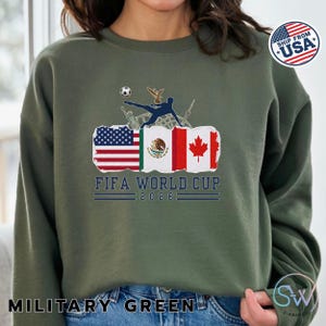 May include: Military green sweatshirt featuring a graphic for the FIFA World Cup 2026. The design includes flags of the USA, Mexico, and Canada, along with a soccer player kicking a ball. The text "FIFA WORLD CUP 2026" is displayed below the flags.