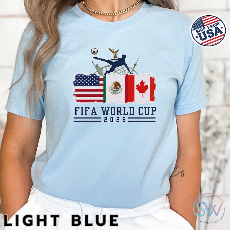 May include: Light blue t-shirt with a FIFA World Cup 2026 graphic. The design includes flags of the USA, Mexico, and Canada, a soccer ball, and a silhouette of a soccer player kicking the ball. The shirt also has the text "LIGHT BLUE".