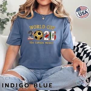 May include: Indigo blue t-shirt with "WORLD CUP 2026" text and a soccer ball graphic. The numbers 2 and 6 are designed with the USA and Mexican flags. The text "USA CANADA MEXICO" is below the graphic.