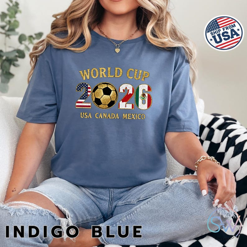 May include: Indigo blue t-shirt with "WORLD CUP 2026" text and a soccer ball graphic. The numbers 2 and 6 are designed with the USA and Mexican flags. The text "USA CANADA MEXICO" is below the graphic.