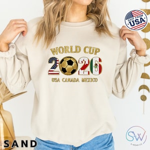 May include: Sand-colored sweatshirt with the text "WORLD CUP 2026" in gold lettering. The number "2" is designed with the American flag, and the "6" with the Mexican flag. A soccer ball is in the center. The text "USA CANADA MEXICO" is below.