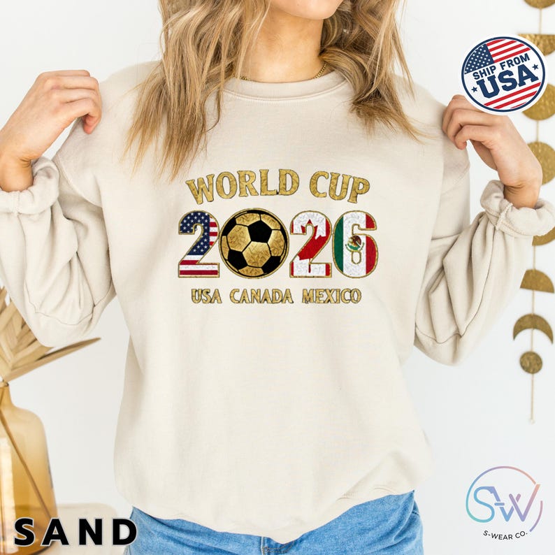 May include: Sand-colored sweatshirt with the text "WORLD CUP 2026" in gold lettering. The number "2" is designed with the American flag, and the "6" with the Mexican flag. A soccer ball is in the center. The text "USA CANADA MEXICO" is below.