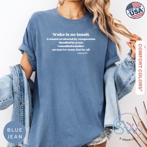 May include: Blue jean Comfort Colors t-shirt with the text "Woke is no insult." The text continues with "It means awakened by compassion. Humbled by grace. Committed to justice - not just for some, but for all." The shirt has rolled-up sleeves.