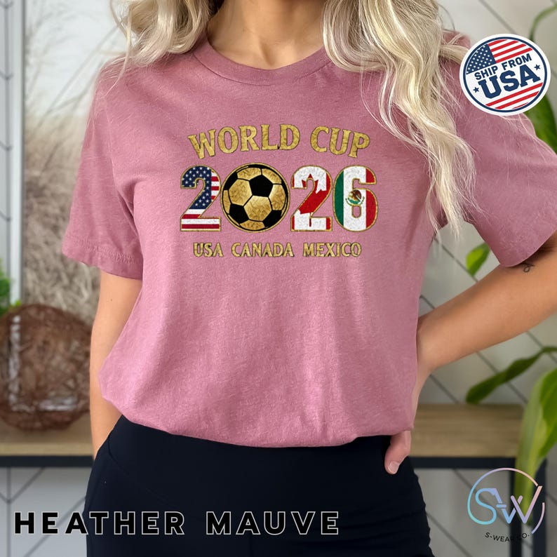 May include: Heather mauve t-shirt with "World Cup 2026" graphic. The design features a soccer ball and the flags of the USA, Canada, and Mexico. The text "USA CANADA MEXICO" is below the numbers. The shirt has short sleeves.