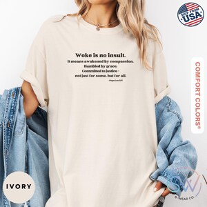May include: Ivory-colored t-shirt with the text "Woke is no insult" and a quote by Pope Leo XIV. The shirt is a Comfort Colors brand and has a relaxed fit. A denim jacket is draped over the arm.