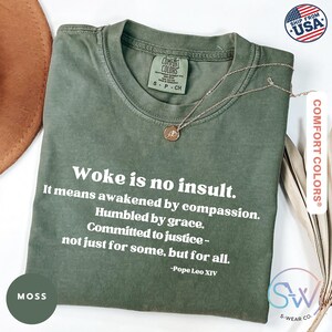 May include: Moss green t-shirt with the text "Woke is no insult." and a quote by Pope Leo XIV. A gold-colored necklace with a pendant is on the shirt. The shirt is from Comfort Colors.