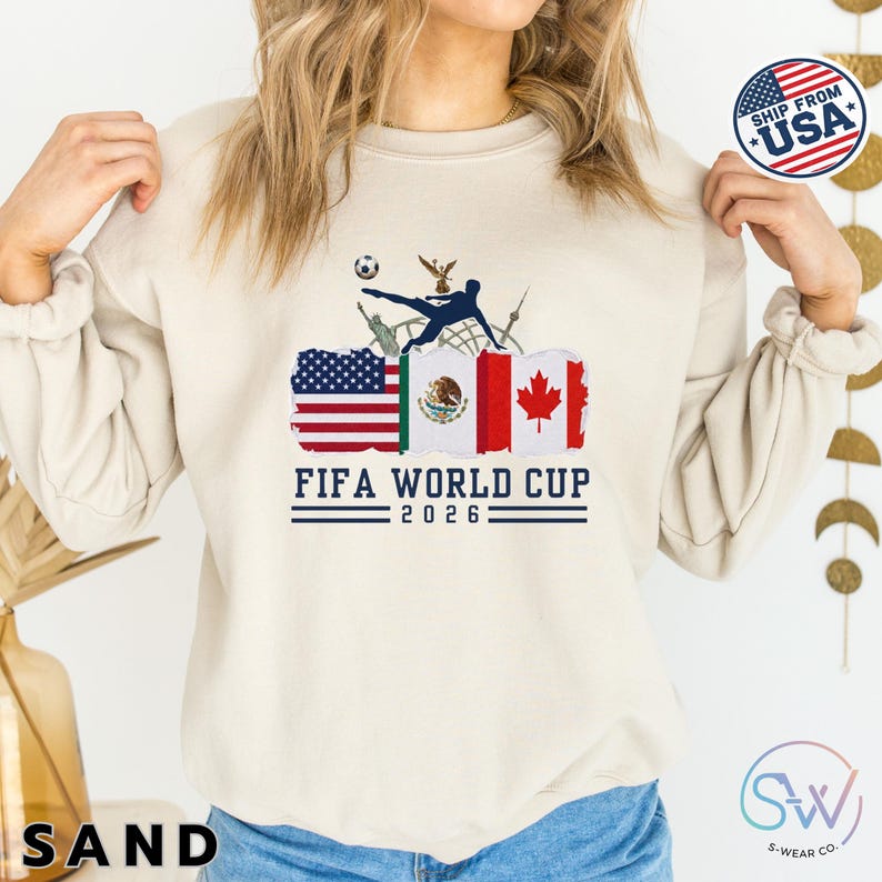 May include: Sand-colored sweatshirt featuring the text "FIFA WORLD CUP 2026" with flags of the USA, Mexico, and Canada. A soccer player silhouette kicks a ball above the flags. The sweatshirt has long sleeves.