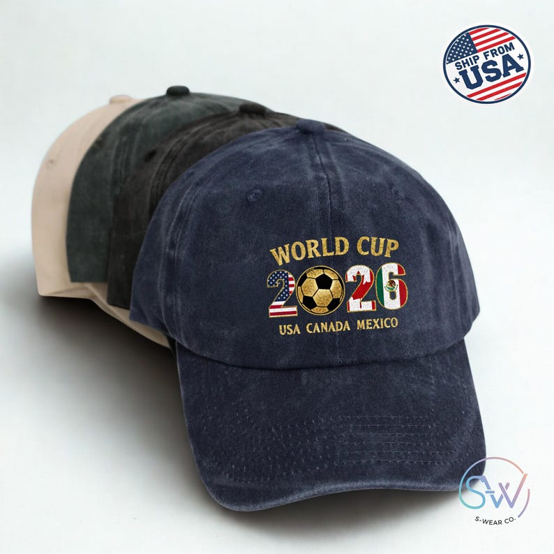 May include: A selection of baseball caps in shades of beige, dark green, and navy. The navy cap displays "WORLD CUP 2026" with a football graphic and the flags of the USA and Mexico. The caps are stacked.