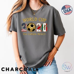 May include: Charcoal gray t-shirt with "World Cup 2026" in gold lettering. The design includes a soccer ball and flags of the USA, Canada, and Mexico. The shirt has a relaxed fit and short sleeves.
