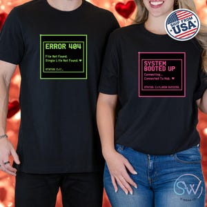 May include: Black t-shirts with computer-themed graphics. One shirt displays "ERROR 404" with text, the other says "SYSTEM BOOTED UP" with text. Both shirts have a square border around the text. The shirts are worn by two people.