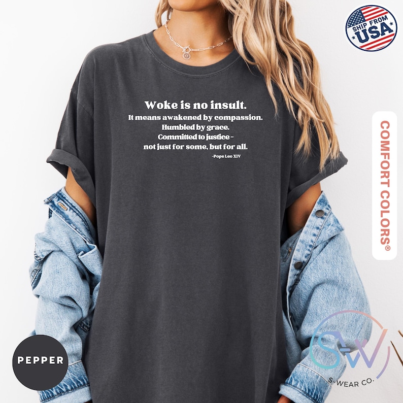 May include: Pepper-colored Comfort Colors t-shirt with the text "Woke is no insult." in white. The text continues with a quote about compassion and justice. The shirt has short sleeves and a relaxed fit. The image also shows a denim jacket.