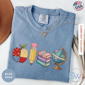 May include: Blue jean Comfort Colors t-shirt with an apple, pencil, books, and globe design. The shirt has a crew neck and short sleeves. The graphic is colorful and features a patchwork style. The shirt is labeled "Comfort Colors" and "Ship From USA".