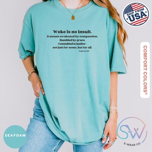 May include: Seafoam green t-shirt with the text "Woke is no insult." and a quote by Pope Leo XIV. The shirt is a Comfort Colors brand and features a relaxed fit. The image also includes a "Ship From USA" sticker.