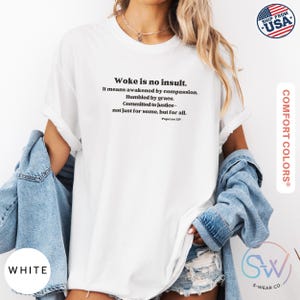 May include: White t-shirt with the text "Woke is no insult. It means awakened by compassion. Humbled by grace. Committed to justice - not just for some, but for all." The shirt is from Comfort Colors and has a "Ship from USA" graphic.