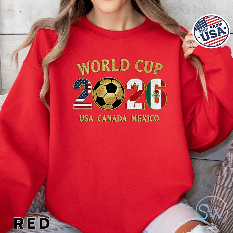 May include: Red sweatshirt with gold text reading "WORLD CUP 2026" and "USA CANADA MEXICO". The number "2026" is designed with the flags of the USA, Canada, and Mexico, and a soccer ball. A "SHIP FROM USA" sticker is visible.