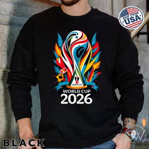May include: A black long-sleeve sweatshirt featuring a colorful graphic of the World Cup 2026 trophy. The design includes abstract shapes in red, blue, yellow, and green, a soccer ball, and the text World Cup 2026 printed in white below the trophy.