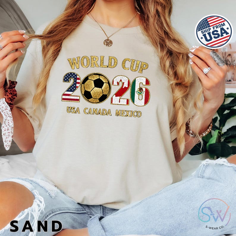May include: A sand-colored t-shirt with the text "WORLD CUP 2026 USA CANADA MEXICO" in gold and patriotic colors. The design includes a soccer ball and the numbers 2026, with the American, Canadian, and Mexican flags incorporated.