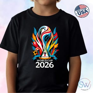May include: A black short-sleeved t-shirt featuring a colorful graphic of the World Cup 2026 trophy. The design includes abstract red, blue, and yellow shapes, a soccer ball, and the text World Cup 2026 printed in white below the trophy image.
