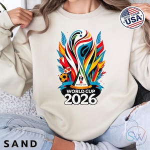 May include: A sand-colored crewneck sweatshirt featuring a colorful graphic of the World Cup 2026 trophy. The design includes abstract red, blue, and yellow shapes, a soccer ball, and the text "WORLD CUP 2026" printed in bold black letters.