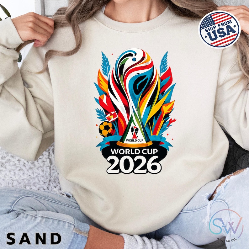 May include: A sand-colored crewneck sweatshirt featuring a colorful graphic of the World Cup 2026 trophy. The design includes abstract red, blue, and yellow shapes, a soccer ball, and the text "WORLD CUP 2026" printed in bold black letters.