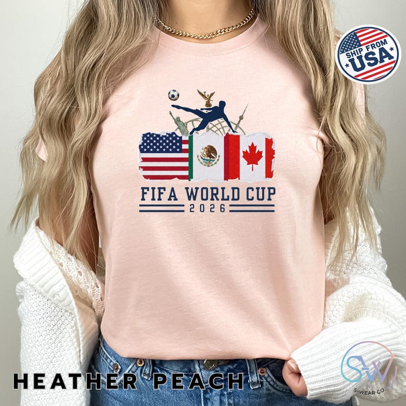 May include: Heather peach t-shirt with a graphic design for the FIFA World Cup 2026. The design includes the flags of the USA, Mexico, and Canada, along with a soccer ball and a player kicking the ball. The text "FIFA WORLD CUP 2026" is printed below the flags.