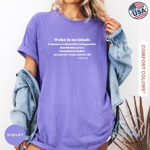 May include: A violet Comfort Colors t-shirt with the text "Woke is no insult" and a quote by Pope Leo XIII. The shirt has short sleeves and a relaxed fit. A denim jacket is draped over the shoulders.