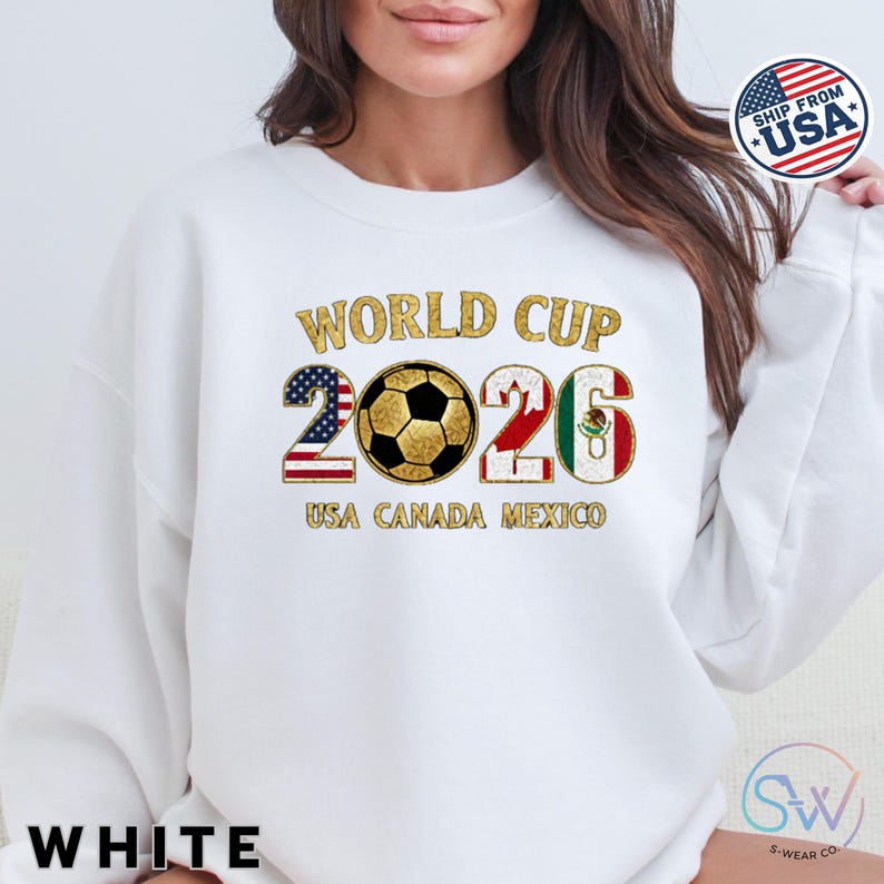 May include: White sweatshirt with "WORLD CUP 2026" in gold glitter lettering. The numbers 2 and 6 are designed with the American and Mexican flags, respectively. A soccer ball is in the center. The text "USA CANADA MEXICO" is below the numbers.