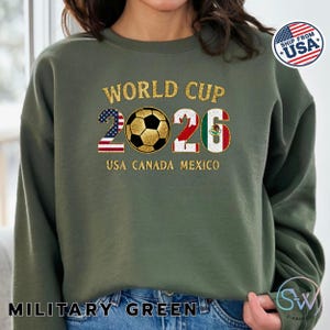 May include: Military green sweatshirt with "WORLD CUP 2026" in gold lettering. The "2026" is designed with the USA and Mexican flags, and a soccer ball. The text "USA CANADA MEXICO" is below the numbers.