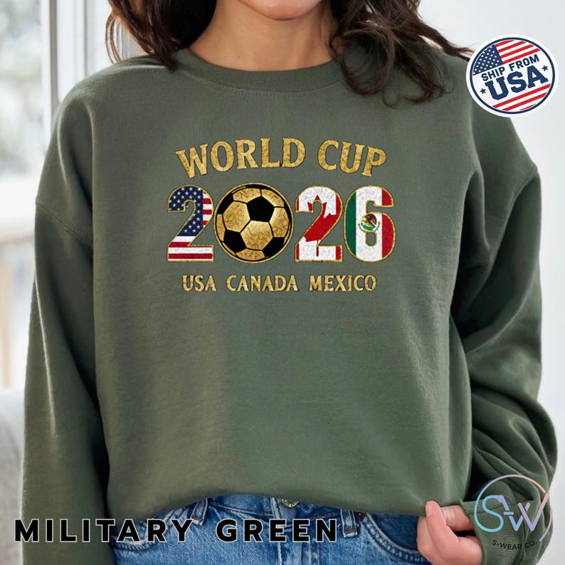 May include: Military green sweatshirt with "WORLD CUP 2026" in gold lettering. The "2026" is designed with the USA and Mexican flags, and a soccer ball. The text "USA CANADA MEXICO" is below the numbers.